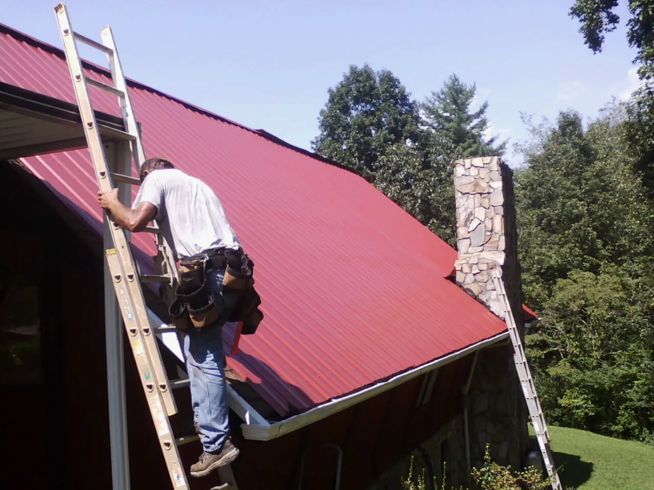 Roofer on a ladder beside a red metal roof and stone chimney — Soffit & Fascia Repair by Roofing Brothers in Wylie